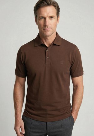 Mann trägt ein braunes kurzärmeliges Poloshirt und dunkelgraue Hose, steht vor einem schlichten hellen Hintergrund.