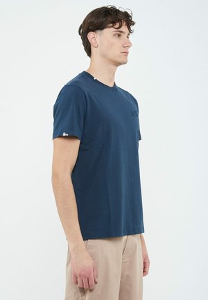 Giovane uomo con capelli corti e ricci, indossa una maglietta blu navy e pantaloni beige, in piedi di lato su uno sfondo bianco.