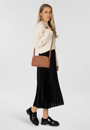 Femme en pull crème et jupe plissée noire portant des chaussures à plateforme noires et un sac bandoulière marron, debout devant un fond uni.