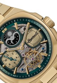 Goudkleurig automatisch horloge met een groene bezel, dubbele maanfase sub-dials, skeletontwerp en gouden accenten op een gestructureerde metalen armband.
