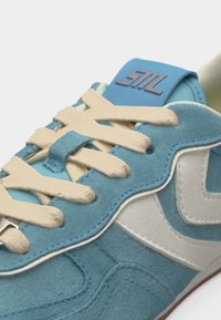 Μπλε και λευκό σουέντ αθλητικό παπούτσι με κρεμ κορδόνια, κοντινό πλάνο στο δέσιμο και στην υφή του υλικού, με λογότυπο σε μπλε ταμπελάκι στη γλώσσα.