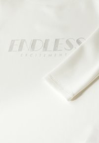 Weißes Langarmshirt mit silbernerprägtem Text "ENDLESS EXCITEMENT", mit glatter Textur und einfachem Rundhals.