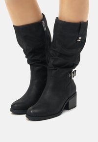 Botas negras de estilo de cuero sintético hasta la rodilla con un diseño holgado, cremallera lateral, acentos en hardware plateado y tacón cuadrado. Acabado texturizado.