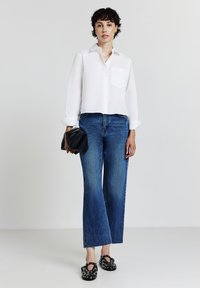Femme portant une chemise blanche à boutons, un jean taille haute bleu, tenant une pochette noire et des ballerines noires à clous, se tenant sur un sol blanc.