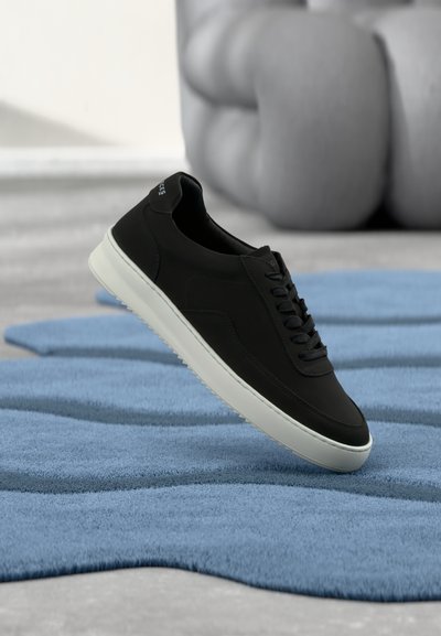 Black low-top sneaker with white sole positioned on wavy blue rug, blurred tufted beige sofa in background.