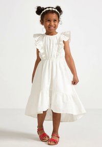 Rochie albă din bumbac cu mâneci cu volane, talie elastică și fustă cu straturi. Purtată cu sandale roșii. Copil zâmbind pe un fundal simplu.