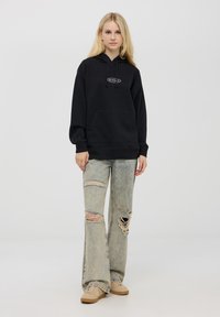 Czarna bluza z kapturem z kieszenią z przodu, zestawiona z jasnoniebieskimi spodniami dzwonami z przetarciami. Beżowe sneakersy dopełniają całość.