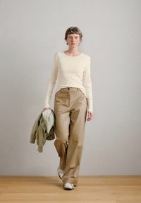 Κρεμ μπλούζα με μακρύ μανίκι, μπεζ παντελόνια σε φαρδιά γραμμή και ανοιχτό πράσινο μπουφάν κρατημένο στο αριστερό χέρι. Λευκά sneakers. Ουδέτερο φόντο με ξύλινο δάπεδο.