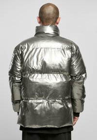 Veste matelassée argentée métallique avec un col haut, des coutures horizontales et une texture brillante. La vue de dos révèle une coupe décontractée et un ourlet bas.
