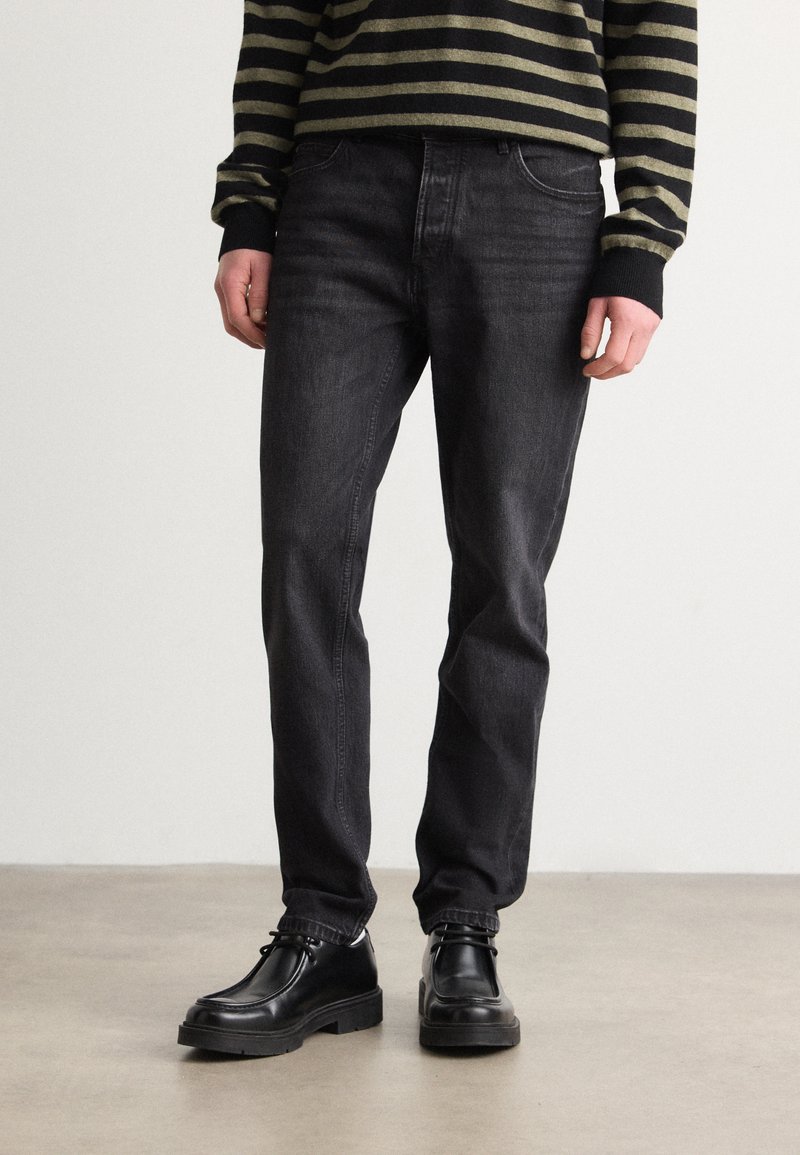 Jeanși din denim negri cu un croi drept, având un design clasic cu cinci buzunare și ușoare decolorări. Purtați cu pantofi negri cu șiret.
