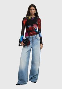 Top negru cu maneci lungi, cu modele abstracte multicolore, purtat cu blugi evazați de culoare albastru deschis și o geantă neagră cu accente roșii.