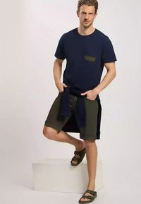 Granatowa koszulka z oliwkową kieszenią, zestawiona z oliwkowymi szortami i ciemnymi sandałami. Model opiera się na białym cokole, prezentując swobodny look.