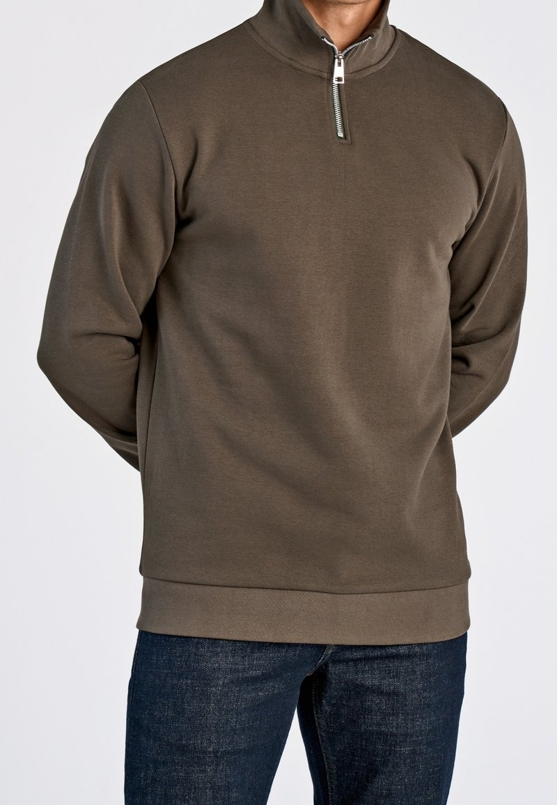 Homme portant un pull marron à demi-fermeture éclair et un jean bleu foncé, debout les mains derrière le dos devant un fond uni.