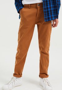 Hnědé bavlněné chinos kalhoty s rovný střih, s zapínáním na knoflík, bočními kapsami a ohrnutými nohavicemi, spárované s bílými teniskami.