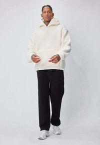 Polaire blanche à capuche avec poche avant, associée à un pantalon noir et des baskets claires ; tissu texturé et coupe décontractée.