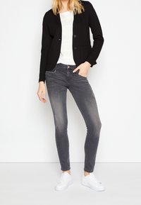 Blazer negro sobre una blusa blanca texturizada, combinado con jeans ajustados grises y zapatillas blancas. El conjunto presenta elementos de diseño mínimos.