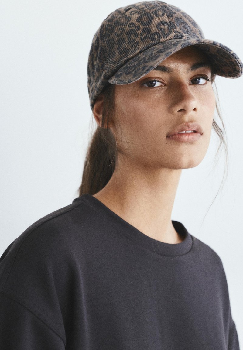 Jeune femme aux longs cheveux foncés portant une casquette à motif léopard et un t-shirt noir à col rond, regardant légèrement sur le côté sur un fond uni.