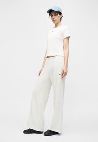 Γυναίκα που φοράει λευκό μπλουζάκι Nike και φαρδιά λευκή παντελόνα με μαύρα αθλητικά παπούτσια και ανοιχτό μπλε καπέλο μπέιζμπολ, στέκεται σε απλό λευκό φόντο.