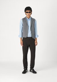 Grå vest over lys blå skjorte, svarte bukser og svarte sneakers med hvite striper. Vestet har knapper og en skreddersydd passform.