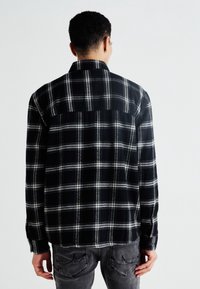 Camicia flanella a quadri bianca e nera con colletto a bottoni, maniche lunghe e orlo leggermente curvo sul retro.