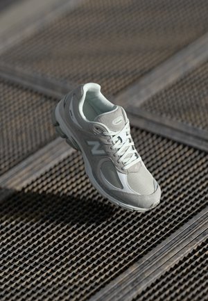 Light gray running shoe with mesh and suede panels, featuring a textured sole and white accents, suspended above a metal grid surface.