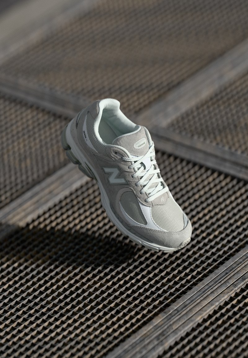 Light gray running shoe with mesh and suede panels, featuring a textured sole and white accents, suspended above a metal grid surface.