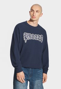 Granatowy sweatshirt wykonany z bawełny, z białym graficznym logo z przodu, okrągłym dekoltem i długimi rękawami. Noszony z niebieskimi dżinsami.
