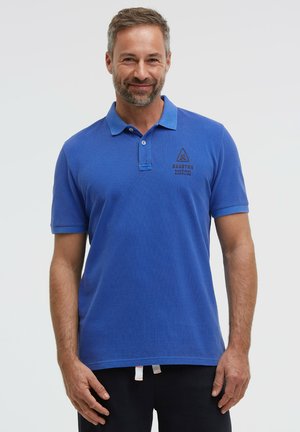 Mann trägt blaues Polohemd mit kurzen Ärmeln und schwarze Hose, steht und lächelt vor einfarbigem Hintergrund.