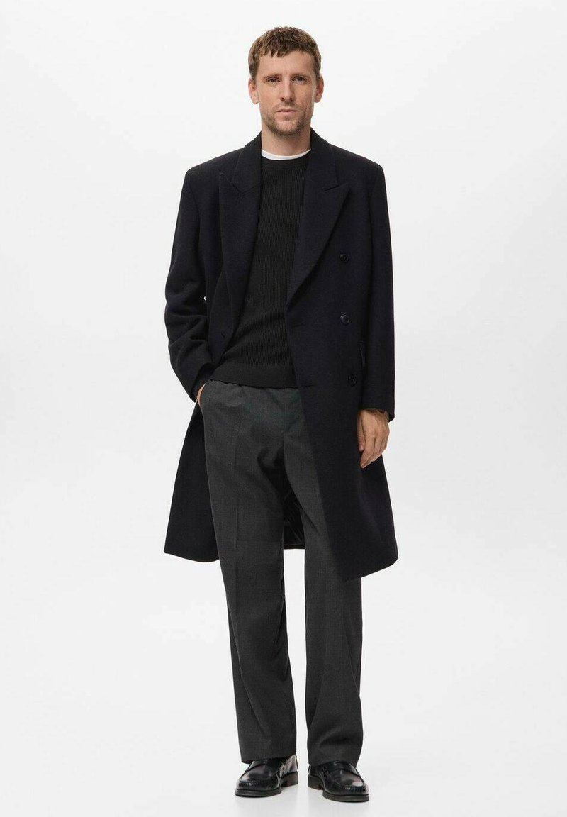 Navy wool overcoat with a double-breasted design, worn over a black knit sweater and gray trousers, paired with black shoes.