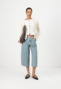 Beige knitted cardigan, white ribbed top with buttons, light blue wide-leg jeans, and black pointed flats with bows, holding a brown clutch.