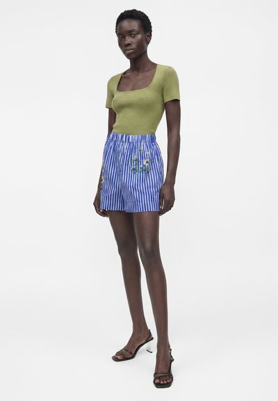 Femme debout portant un haut vert à manches courtes, un short rayé bleu et blanc avec broderie florale, et des sandales à talons noires.