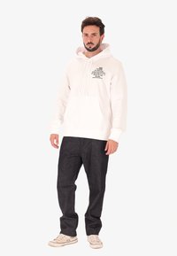 Felpa bianca con tasca frontale, cappuccio regolabile con cordino e testo grafico nero. Abbinata a pantaloni di denim scuri e sneaker bianche.
