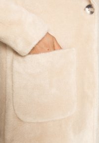 Abrigo de un suave color beige claro y esponjoso, confeccionado con material blando, que presenta un gran bolsillo frontal y un cierre de botón redondo en el lateral.