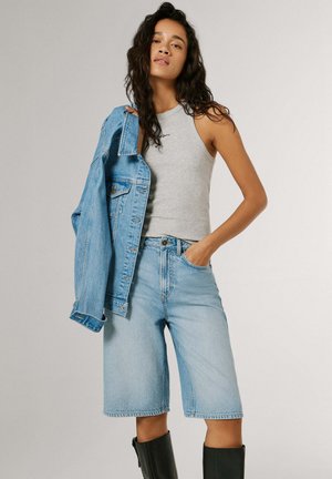 Kvinne iført grå ermeløs topp, lyseblå jeansshorts til knelengde, svarte støvler, og holder en lys blå jeansjakke over den ene skulderen.