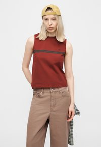 Νεαρή γυναίκα που φοράει ένα μουσταρδί κίτρινο καπέλο προς τα πίσω, αμάνικο κόκκινο τοπ με μαύρες ρίγες, μπεζ παντελόνι και κρατάει ένα καρό πουκάμισο.