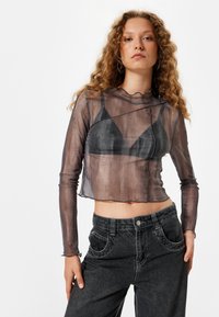 Crop top noir transparent à manches longues, décolleté festonné, design en couches, porté par-dessus un bralette noir, associé à un jean foncé taille haute.