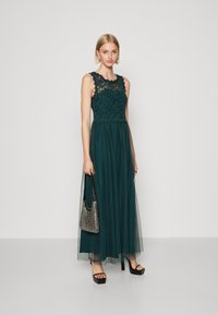 Rochie lungă, de culoare verde închis, cu un corset din dantelă, suprapunere transparentă și talie ajustată. Modelul ține o geantă metalică și poartă sandale cu toc gros.