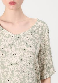 Grønn blomstermønstrete topp laget av lett materiale. Har en V-hals og korte ermer med svarte detaljer gjennom hele designet.