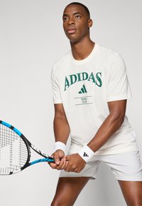 Camiseta branca de manga curta com o logótipo verde "ADIDAS", segurando uma raquete de ténis com um quadro preto e azul. Calções brancos e pulsos.