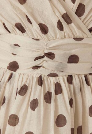 Beige stof met grote bruine stippen, met geplooide stiksels en een gedraaide knoopdetail ter hoogte van de taille van een kledingstuk.