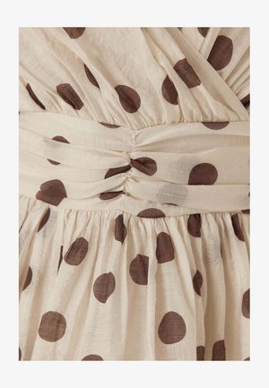 Beige stof met grote bruine stippen, met geplooide stiksels en een gedraaide knoopdetail ter hoogte van de taille van een kledingstuk.