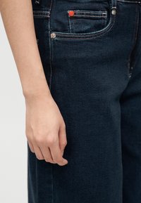 Το αριστερό χέρι ακουμπά δίπλα σε σκούρο μπλε τζιν με ορατές ραφές και μικρή μπροστινή τσέπη με κόκκινη λεπτομέρεια κουμπιού.