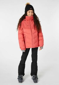 Giacca puffer corallo con colletto alto, accenti grigi nei polsini e due tasche con zip, abbinata a pantaloni neri e scarponi da sci.
