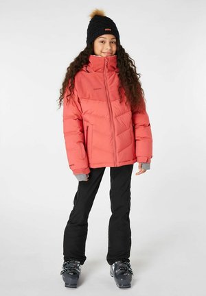 Giacca puffer corallo con colletto alto, accenti grigi nei polsini e due tasche con zip, abbinata a pantaloni neri e scarponi da sci.