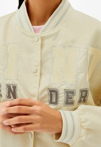 Jasno beżowa kurtka bomberka z prążkowanym kołnierzem, zapięciem na guziki oraz wyrazistymi haftowanymi literami w jasnym żółtym i szarym kolorze z przodu.