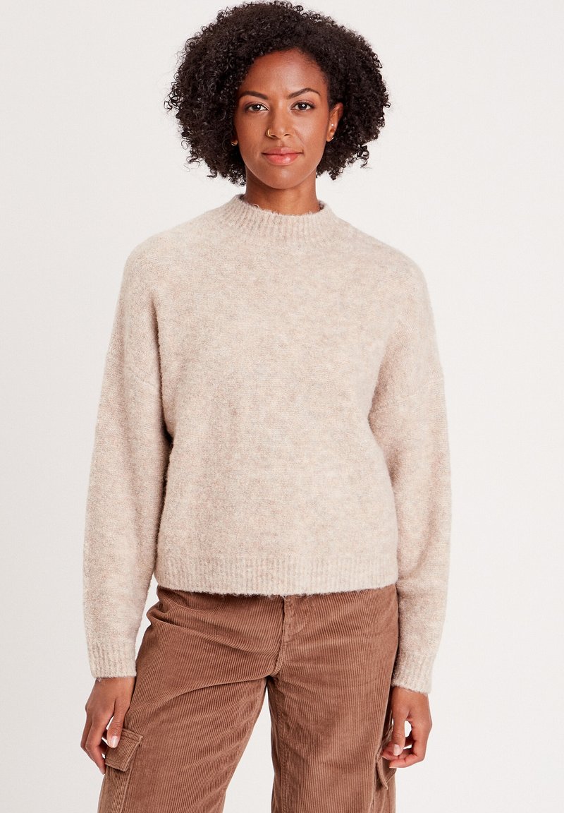 Pull en tricot beige avec un col monté, des manches longues et un ourlet côtelé. Le tissu a une texture douce et pelucheuse avec des variations de motifs subtiles.