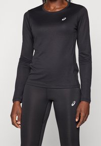 Czarna, długosleeve'owa koszulka sportowa oraz dopasowane legginsy wykonane z gładkiego, elastycznego materiału. Na piersi koszulki znajduje się małe logo.