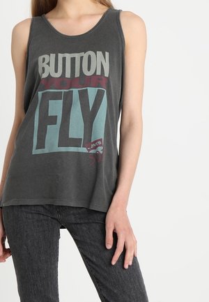 Kobieta ubrana w ciemnoszary tank top bez rękawów z napisem "BUTTON YOUR FLY" dużymi blokowymi literami oraz ciemnoszare dżinsy.