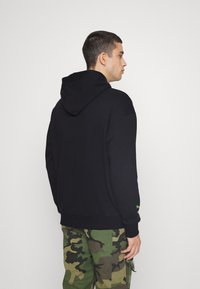 Felpa nera con cappuccio regolabile, polsini e orlo a coste, con un dettaglio verde sulla manica. Abbinata a pantaloni cargo in camouflage.