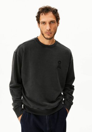 Mann mit kurzem, lockigem Haar und leichtem Bart, trägt ein dunkelgraues Sweatshirt und dunkle Hosen, steht mit den Händen in den Taschen vor weißem Hintergrund.
